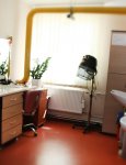 Wnętrze Domu Pomocy Społecznej. Pomieszczenie fryzjera. Wzdłuż ściany stoją meble ze sprzętem fryzjerskim. Przy przeciwległej ścianie dwie umywalki. W tle okno zasłonięte kremową żaluzją pionową